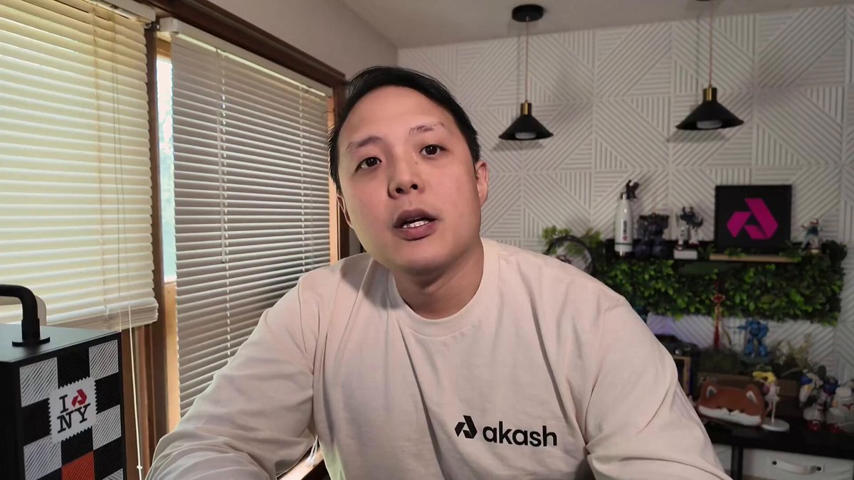 The image shows an Asian man wearing a white shirt, sitting at a desk with a computer monitor behind him. The man appears to be giving a presentation or participating in a video call using a webcam. There are also several potted plants placed around the room, adding some greenery and life to the space.