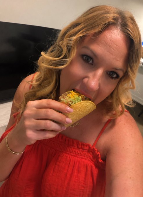 A woman with wavy blonde hair in a red top takes a selfie while biting into a hard-shell taco filled with lettuce, cheese, and other toppings, capturing the perfect "Taco Tuesday Gm" vibe.