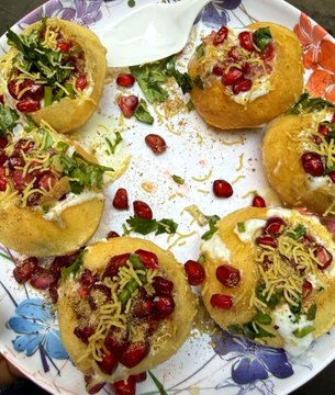 A plate of Indian street food, likely raj kachori or dahi puri, topped with yogurt, pomegranate seeds, sev, and cilantro on a floral-patterned plate.