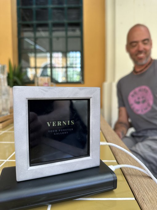 The image features a grey picture frame with a white screen, placed on top of a table. The frame has a black border around its edges. In the background, there are two people sitting at a dining table, one person wearing a pink shirt and another in a gray shirt. A potted plant is also visible near them. The image seems to be related to an art-related event or gathering, possibly a gallery opening or exhibition.
