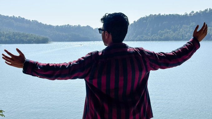 A man wearing sunglasses and a red and black striped shirt stands with his back to the camera, arms outstretched toward a scenic lake surrounded by forested hills.