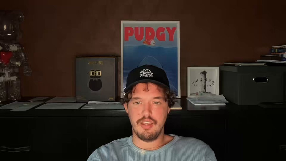 The image shows a man wearing a black baseball cap sitting at a desk with a computer monitor, possibly participating in an online meeting or video call. The background features a brown wall with various posters hanging on it, including one that says "Purggy". There are also several books and papers scattered around the room, suggesting a workspace or study area.