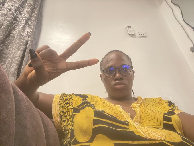 A person wearing glasses and a yellow patterned top makes a peace sign at the camera in a casual indoor selfie.