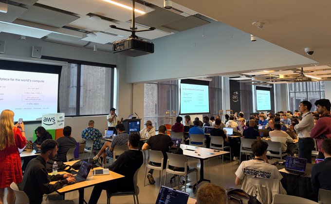 A large conference room or classroom with many attendees working on laptops, with AWS signage and presentation slides visible on screens.