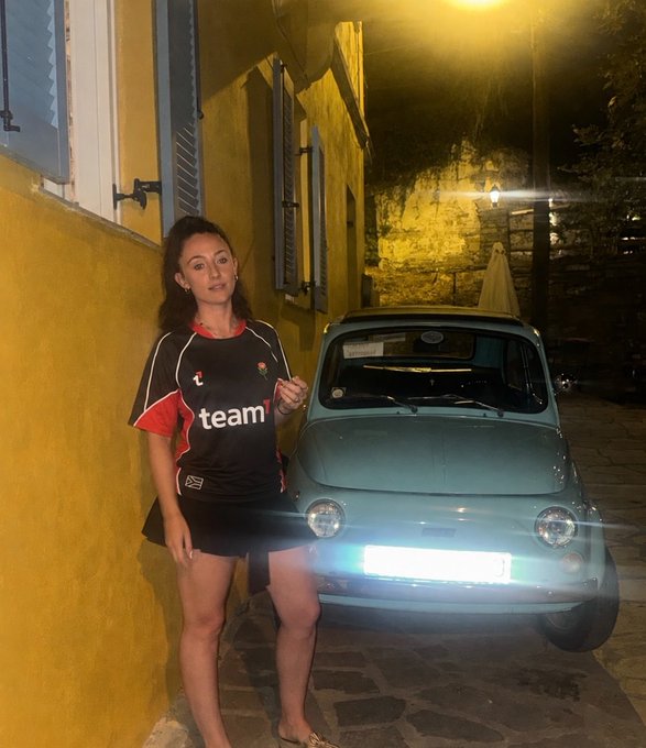 A woman in a black "team" jersey and shorts stands next to a light blue classic car with its headlights on, parked on a cobblestone street at night.