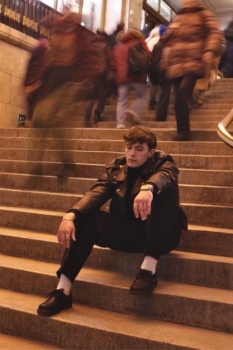 A young man wearing a black leather jacket, dark pants, and white socks sits alone on stone steps while blurred figures of people rush past him going up the stairs, creating a sense of motion and isolation.