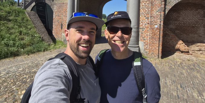 A selfie of two smiling men in baseball caps and sunglasses standing on a cobblestone path in front of a historic brick archway, both wearing backpacks.