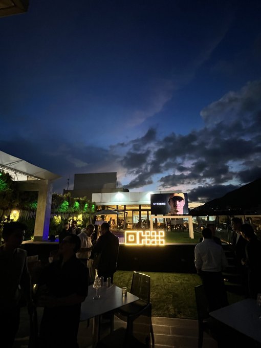 An outdoor evening event features a large screen displaying a 'MY HERO' portrait, with a glowing cube sign and guests gathered at tables.