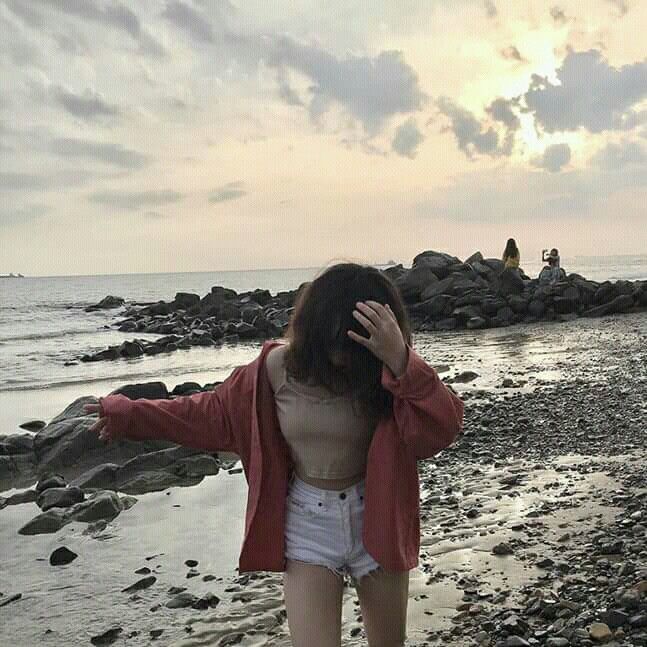 The image features a woman standing at the edge of a rocky beach with her hair blowing in the wind, wearing shorts and a red jacket. She appears to be posing for a photo or enjoying the view. The background shows the ocean meeting the sky, creating an interesting contrast between the natural elements. The post about @quipnetwork is located near the woman's position on the beach, making it easy to connect her presence with the content of the image.