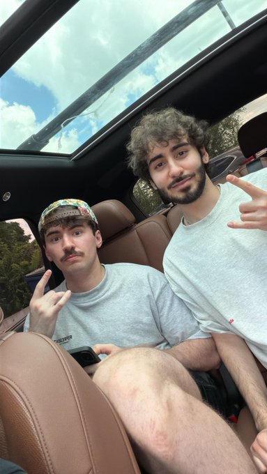 Two young men are taking a selfie inside a car with a panoramic sunroof, both wearing gray t-shirts and making hand gestures toward the camera, with one wearing a colorful patterned headband. The casual, energetic vibe fits the post's theme of friends hyped up and creating $ANSEM content together as a content-creating duo.