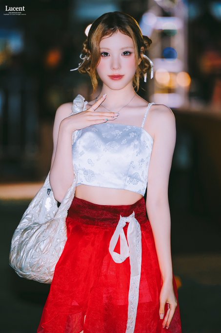 A digitally rendered illustration of a young woman with hair in twin buns, wearing a white floral top and red skirt with a white sash, holding a silver bag against a nighttime city background, labeled 'Lucent' in the corner.