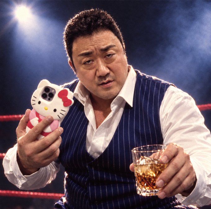A man in a blue pinstripe vest and white shirt holds up a Hello Kitty phone case while sitting in what appears to be a wrestling ring, holding a glass of amber liquor.