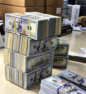 Stacks of $100 bills bundled with currency straps reading "$10,000" are piled on a reflective surface, visually representing the "$1M" referenced in the post. The image directly corresponds to the caption's choice between taking the cash or spending time with CZ (Binance's founder Changpeng Zhao).