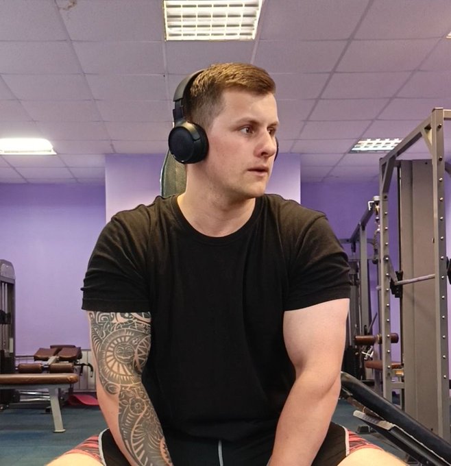 A muscular man with a tattoo on his right arm, wearing a black shirt and headphones, sitting in a gym.
