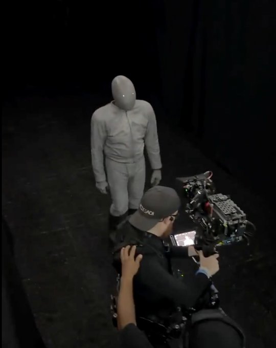 Behind-the-scenes view of a film production featuring a person in a grey full-body suit and mask standing in a dark set, while a camera operator films them.