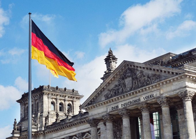 The German flag flying at half-mast in front of the Reichstag building in Berlin, with the inscription 'DEM DEUTSCHEN VOLKE' visible on the pediment.