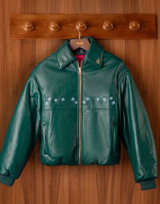 A glossy emerald green leather bomber jacket with floral embroidery hangs on a wooden coat rack with brass hooks; the hanger reads "Made."