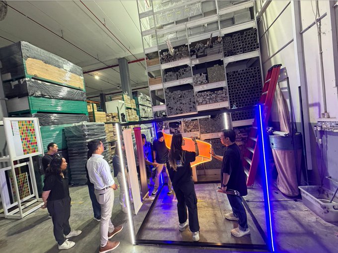 A group of people in a warehouse are prepping an installation, with one person holding an illuminated orange arrow and another holding a glowing blue light bar, while shelves of metal pipes and stacked palletized goods surround them in preparation for #SuiBasecamp at #Token2049 in Singapore.
