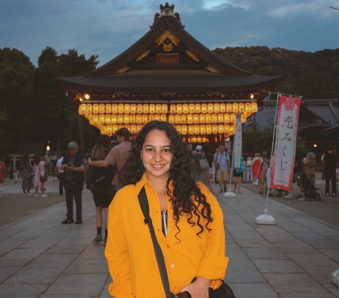 A woman with long curly hair wearing a yellow shirt smiles in front of a Japanese shrine illuminated with lanterns, with a banner displaying the characters '恋みくじ' visible to the right.