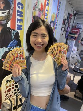 A smiling young woman holding several promotional cards or flyers, standing in front of posters with 'GHEN' text and motorcycle imagery.