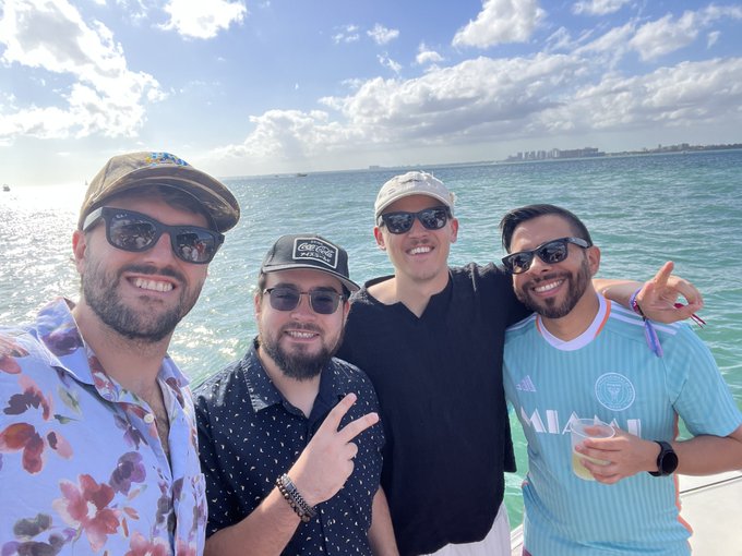 Four smiling friends pose for a selfie on a boat, with the turquoise ocean and a distant city skyline visible behind them.