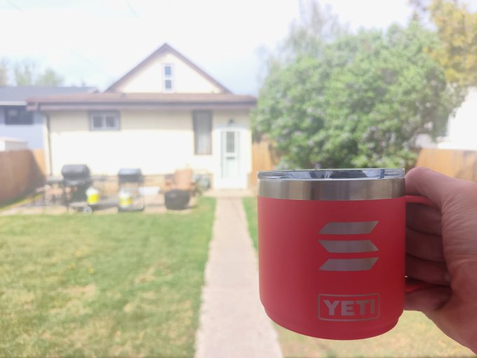 The image shows a person holding up a red mug with the letters "S" and "B" printed on it, which appears to be a coffee cup or thermos. The background of the photo features a house and a backyard with chairs and grills visible outside. The text in the post reads "GM Solana".