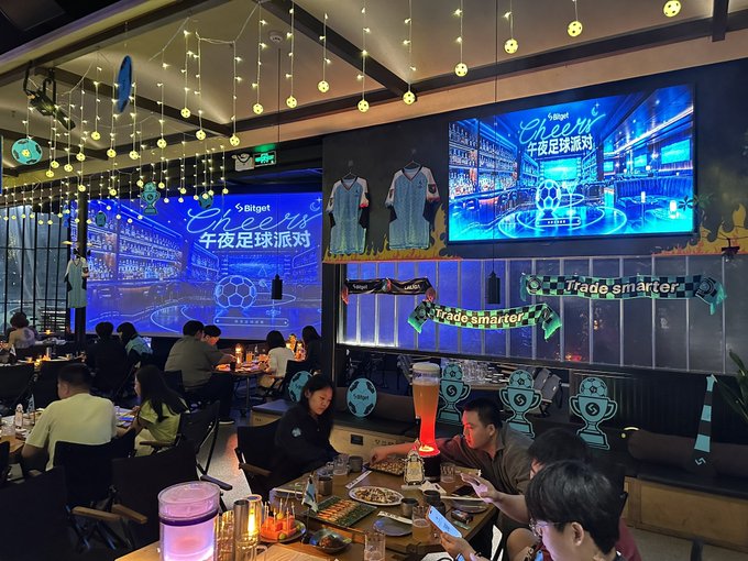 A bustling bar/restaurant scene decorated for a 'Bitget Cheers' football viewing party (午夜足球派对), with large screens displaying the event, football jerseys on display, 'Trade smarter' banners, and patrons seated at tables.