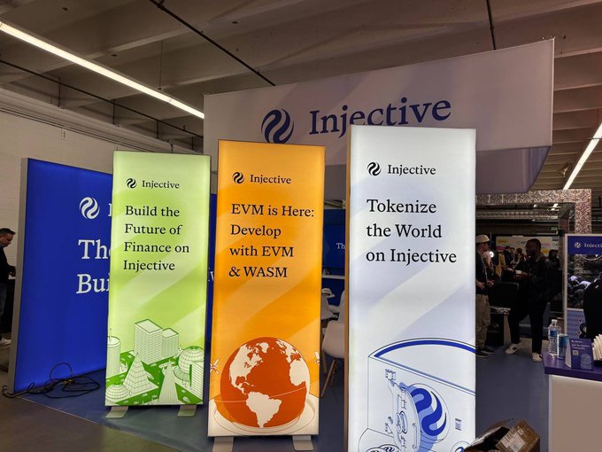 Three vertical banners at an Injective blockchain booth display messages including "Build the Future of Finance on Injective," "EVM is Here: Develop with EVM & WASM," and "Tokenize the World on Injective."