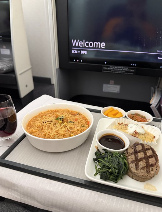 An in-flight meal is served on a tray featuring a bowl of ramen noodles, a steak with grilled vegetables, and side dishes, with a screen displaying "Welcome ICN ➝ DPS" indicating a flight from Incheon to Denpasar. The image reflects the travel portion of the post, capturing the journey en route to Bali for the Alpha Arena Grand Final.