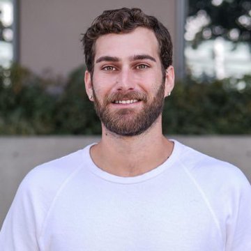 A smiling man with curly brown hair and a beard, identified as Zak Folkman, co-founder of World Liberty Financial, wearing a white crew-neck shirt outdoors with greenery in the background.