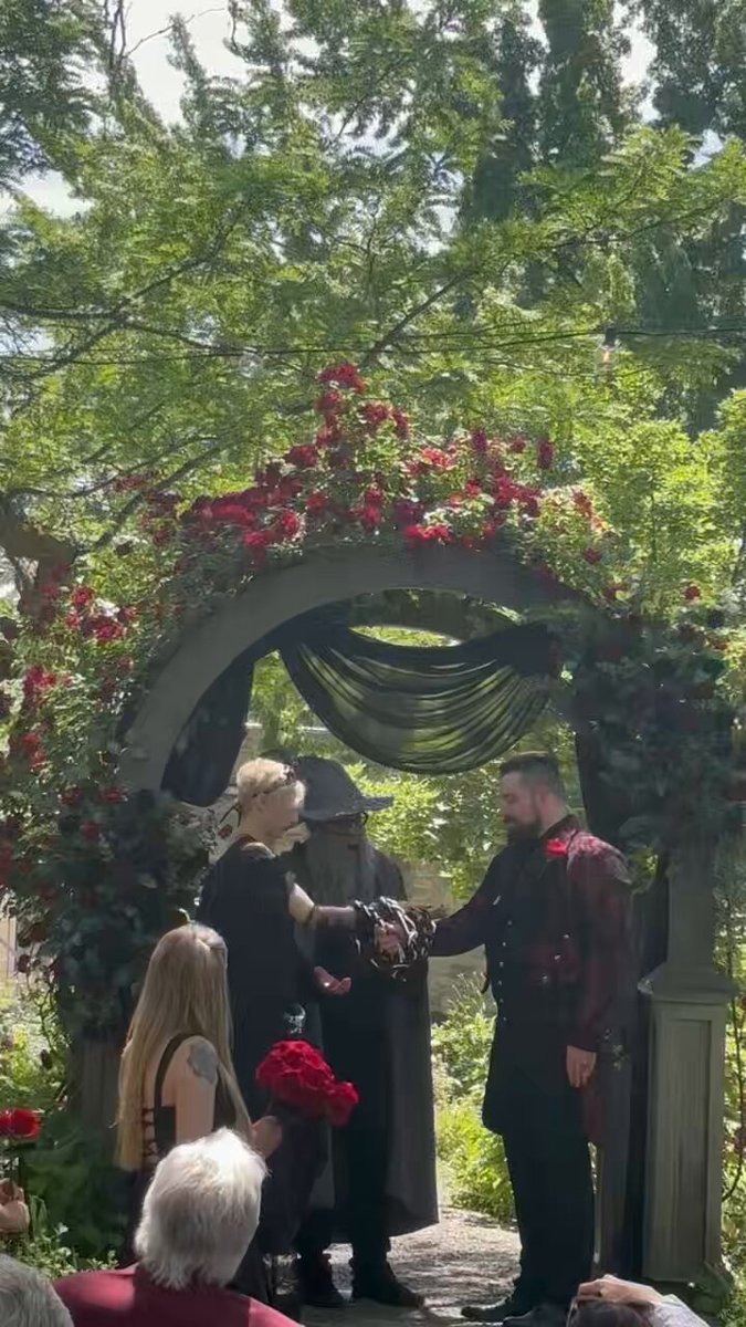 The image shows two people dressed in medieval clothing, standing under an archway with red flowers at their wedding ceremony. The couple appears to be engaged in a hand-fasting spell as part of their wedding tradition. A group of people can be seen watching the ceremony from behind them.