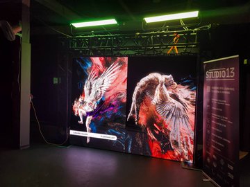 An indoor exhibition or event space showing a large two-panel digital display featuring colorful artwork of bird wings in red, orange, and blue hues, with a vertical banner labeled "STUDIO 13" on the right side.