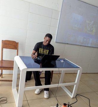A young man sits at a white desk in a classroom, working on a laptop, with a projector screen visible on the wall behind him.