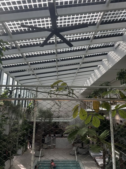 An indoor atrium view looking up at a large industrial ceiling fan under a glass paneled skylight, with tropical plants, a chain-link safety net, and a swimming pool visible below.