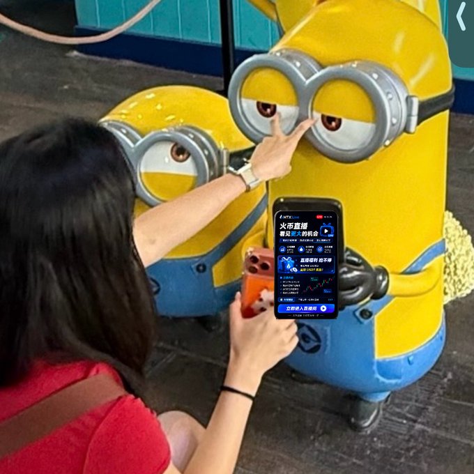 A woman in a red top makes a peace sign while taking a photo with her phone in front of two large Minion statues, with the phone screen displaying a "HTX Live" livestream page offering red envelopes ("红包") and showing a crypto market chart, tying into the caption's playful "I heard you watch HTX_Live too?" reference about catching her tuning into HTX's livestream rewards.