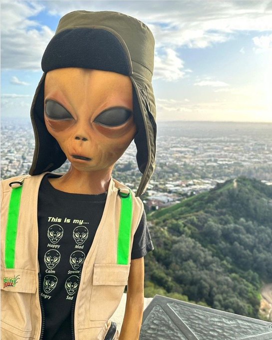 An alien figurine wearing a khaki aviator hat and vest stands at a scenic overlook, wearing a t-shirt displaying various alien expressions with the text "This is my..." followed by labels like "Happy," "Mad," "Calm," and "Stern." The casual "gm ct" greeting pairs with the laid-back, fun vibe of the extraterrestrial tour guide exploring Connecticut.