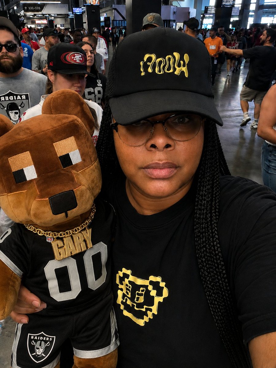 A selfie of a woman wearing glasses and a black cap with gold graffiti-style lettering, holding a plush bear mascot wearing a Raiders jersey labeled 'GARY 00' at what appears to be a sports event.