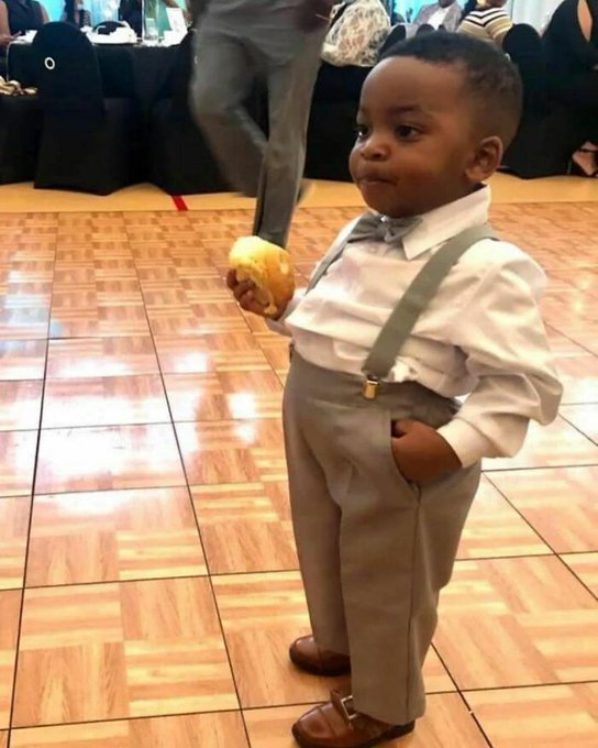 A young child dressed in formal attire with suspenders, white shirt, bow tie, and gray pants, standing on a tiled floor while holding a piece of food.