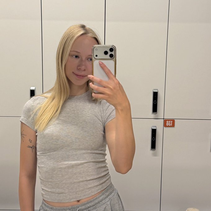 A blonde woman taking a mirror selfie in front of white lockers, wearing a gray fitted t-shirt and sweatpants, holding an iPhone with a white case.