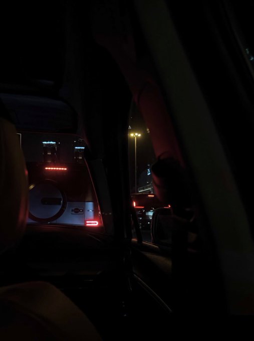 A view from inside a dark vehicle looking out the window at night, with red interior lighting illuminating the back of the front seats, and a streetlamp visible outside.