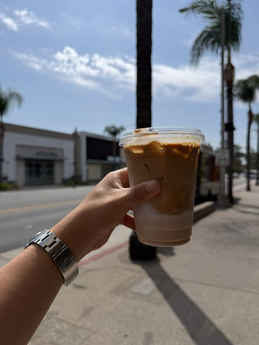 A hand wearing a silver watch holds a plastic cup of iced coffee on a sunny sidewalk lined with palm trees.