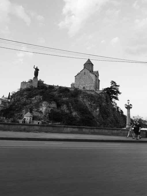 A black and white photograph captures a stone church perched atop a rocky hill, with a large statue of a figure holding a cross standing prominently to the left. The scene is viewed from across a wide, empty road where power lines stretch overhead.