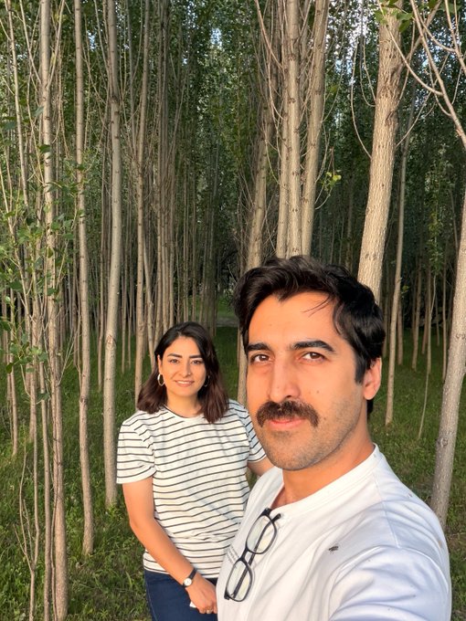 A man and woman take a selfie in a lush grove of tall, slender trees, both smiling at the camera. The outdoor, nature-filled setting perfectly embodies the "touching grass" sentiment of the post, contrasting laid-back real-world time spent outside with online crypto drama.