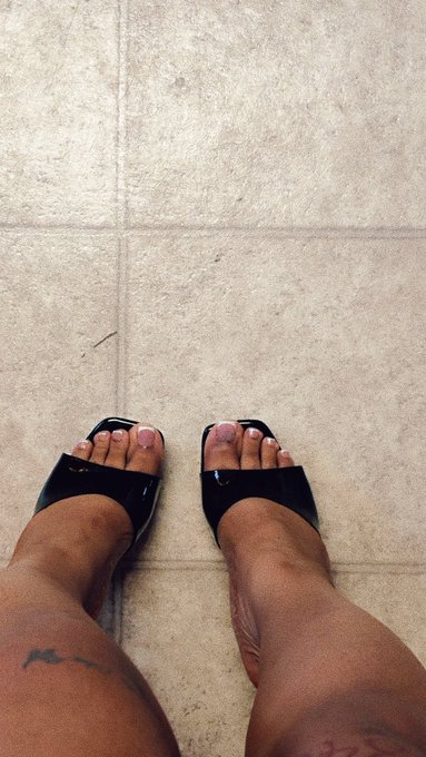 A pair of feet wearing black sandals stands on a beige tiled floor, showcasing a fresh pedicure with light pink polish on the toenails. A tattoo is partially visible on one ankle.