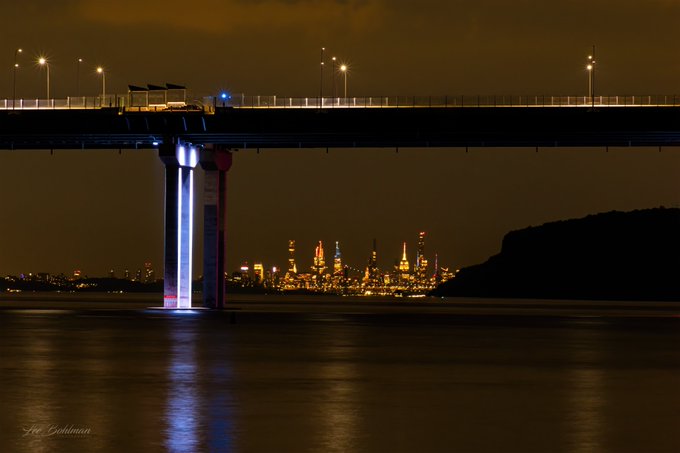 The image shows a bridge over water, with lights illuminating the scene at night. In the background, there are tall buildings that appear to be part of a city skyline. The bridge spans across the body of water below, connecting two points separated by the water. The lights on the bridge and in the surrounding area create a beautiful contrast against the dark sky, highlighting the architectural details of both the bridge and the buildings in the background.
