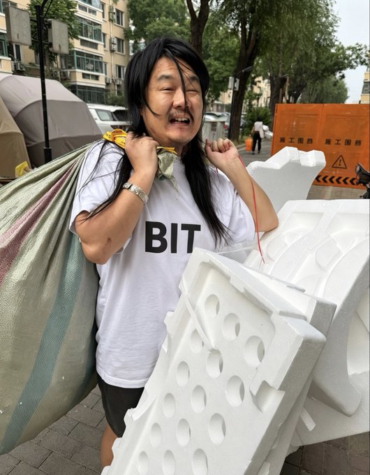 A person with long black hair, wearing a "BIT" t-shirt, struggles under the weight of a large white styrofoam mold and a striped bag, with construction barriers labeled "施工围挡 围工围挡" visible in the background. The image humorously captures the exhaustion of learning complex options trading in the US stock market, visually representing the overwhelming mental and physical toll of tackling a new, challenging skill.