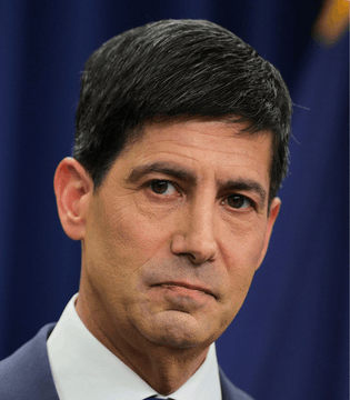 A close-up portrait of a man in a dark suit with short dark hair against a blue background.