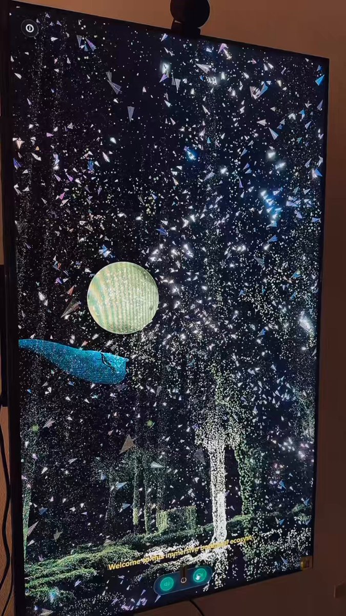 The image shows a large flat screen TV mounted on the wall, displaying an interesting scene of stars and planets. The TV is turned off, but there are some small lights around it that might be used for projection or other purposes. In the corner of the room, there is a black speaker positioned to enhance the audio experience while watching this captivating display. 

The post features a photo of a large screen TV mounted on the wall with a caption about Tezos and a 1/1 message. The image also includes a small picture of a tree in front of a house, which adds an interesting detail to the scene displayed on the television screen.