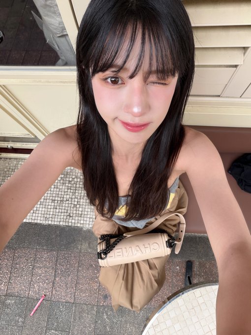 A high-angle selfie of a young Asian woman with long dark hair winking, wearing a gray tube top and a beige Chanel crossbody bag.