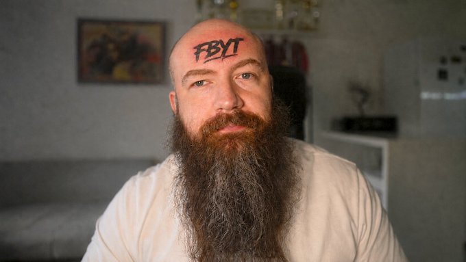 A bald man with a long gray and red beard sits indoors wearing a white t-shirt, with 'FBYT' written across his forehead.